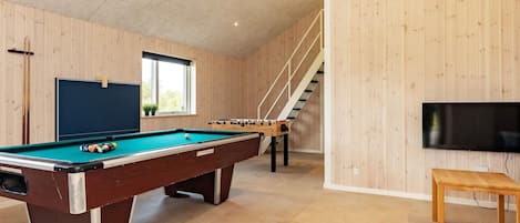 A pool table with a foosball table and a television in a room with wooden walls and a staircase.