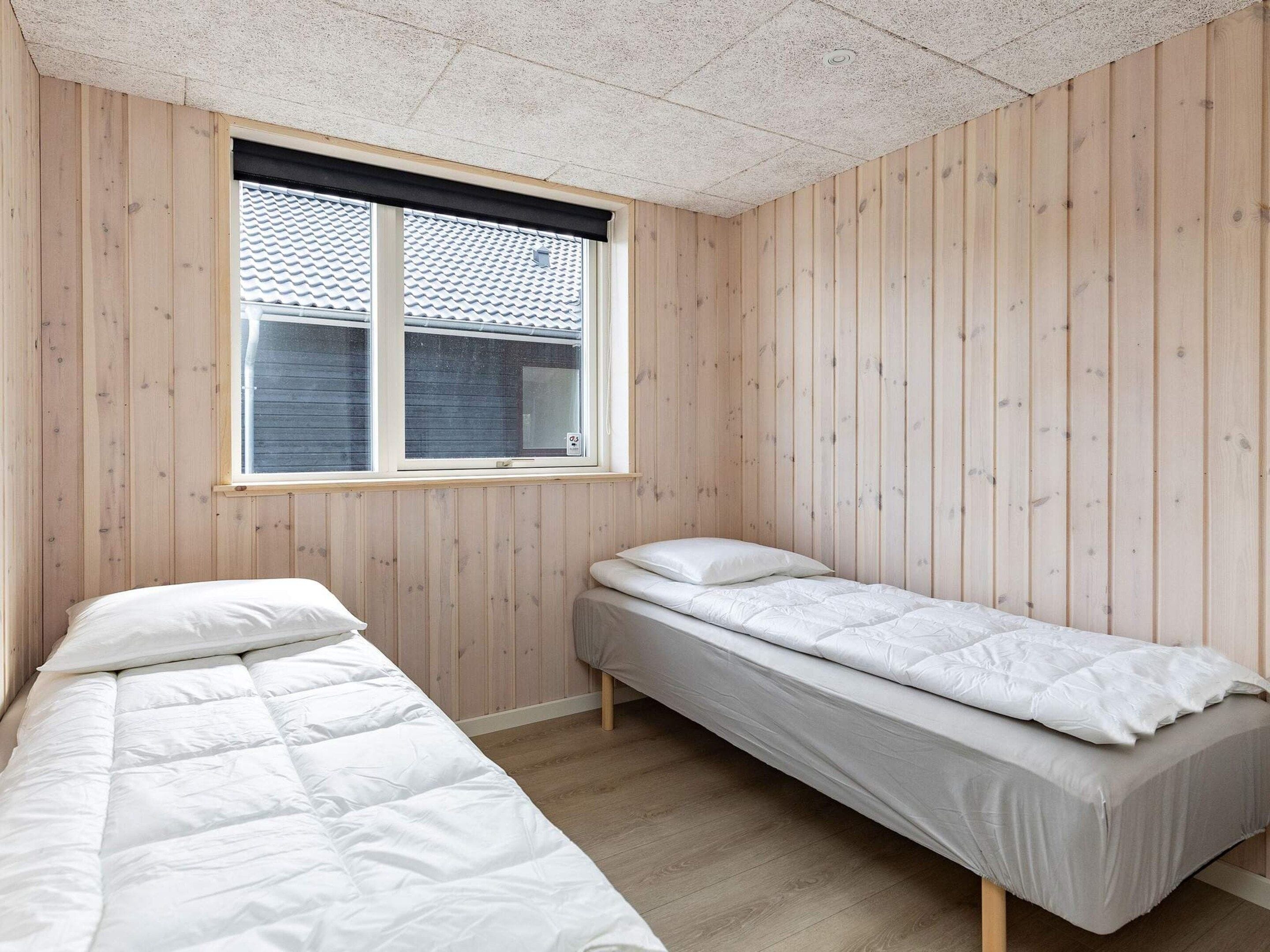 A room with two single beds, wooden walls, a window with blinds, and a ceiling with a textured finish.