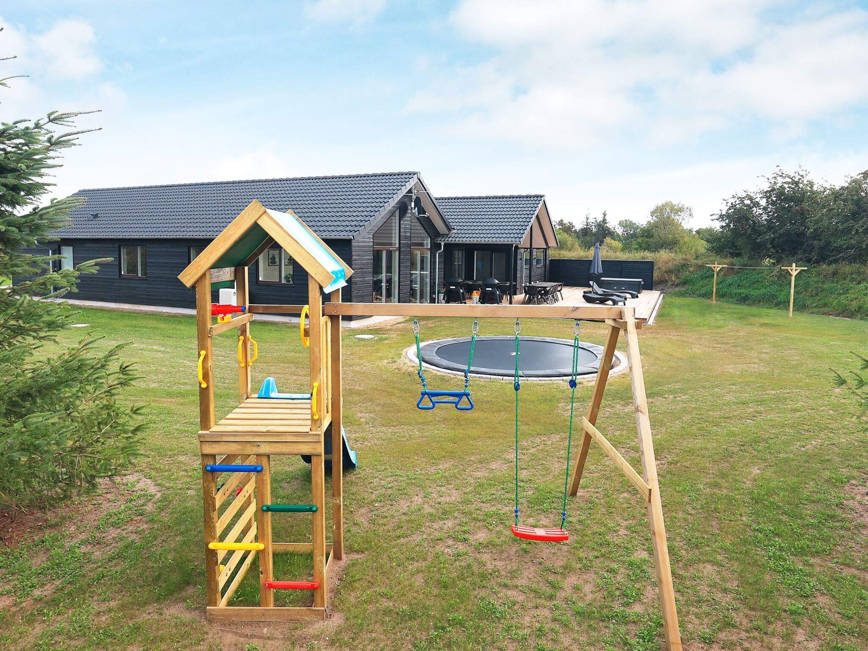 Eine Holzspielanlage mit Rutsche und Kletterelementen, ein Trampolin und im Hintergrund ein Haus.