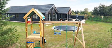 A wooden play structure with a slide and climbing elements, a trampoline, and a house in the background.