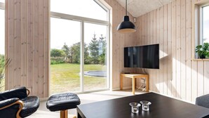 Una sala moderna con un televisor grande, paredes de madera, una mesa de comedor y vistas a un área verde exterior.