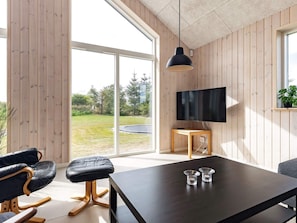 A modern living room with a large TV, wooden walls, a dining table, and a view of a grassy outdoor area. - 20 Person Holiday Home in Hojslev (Hojslev)