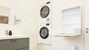 A compact laundry room with two stacked washing machines, a small sink with a soap dispenser, a trash bin, and a white shelf unit.