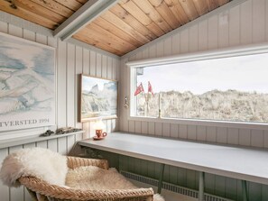 Una habitación con techo alto, un ventanal con vistas a las dunas, una silla de mimbre y dos cuadros enmarcados.