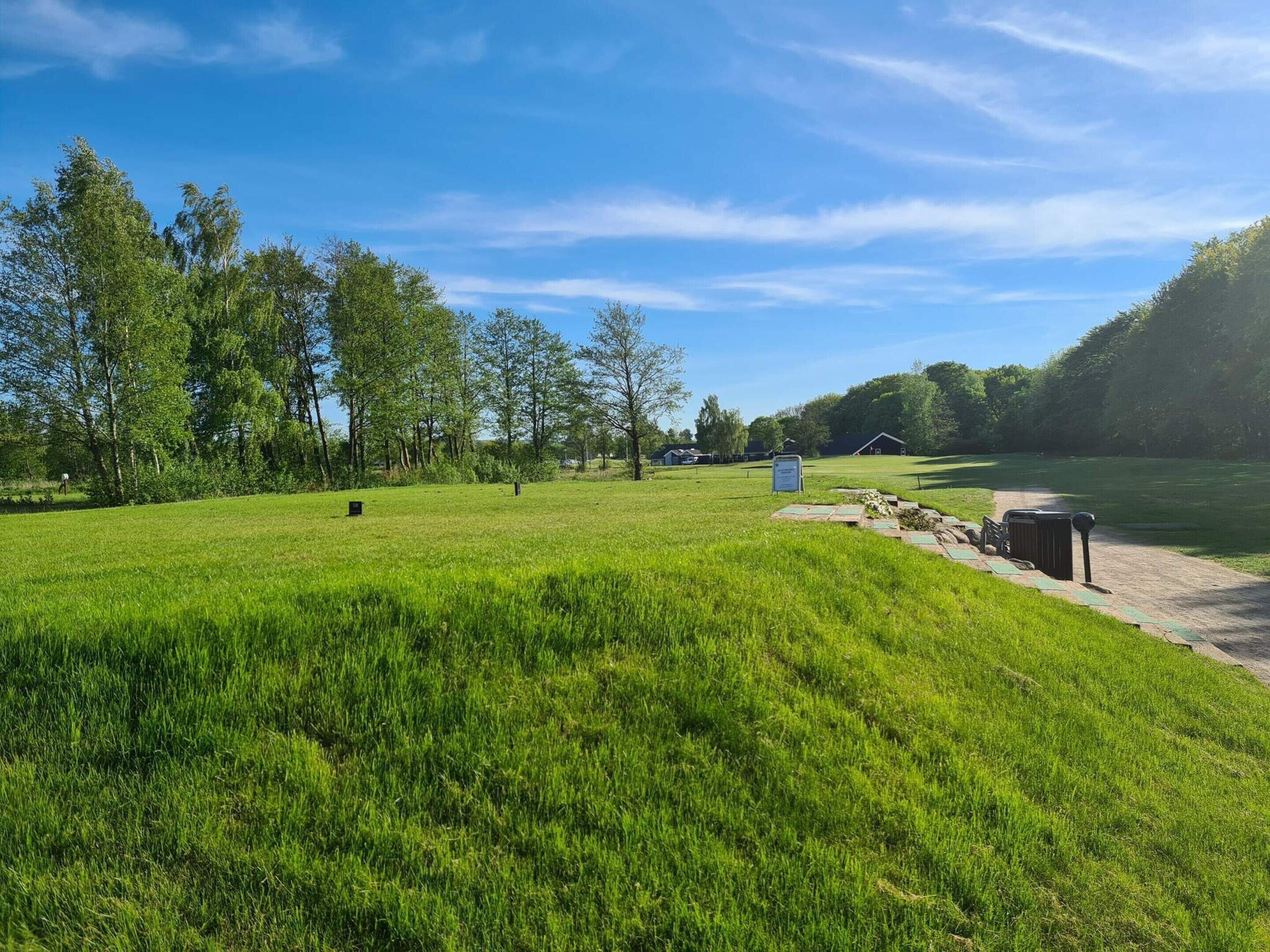 A grassy field with a clear sky, trees, and a few buildings in the distance.