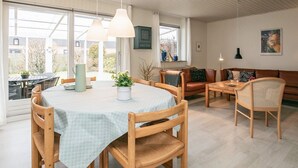 A spacious living area with a round table, wooden chairs, a sofa, and a couch. There are large windows providing natural light and a view of the outdoors. - 6 Person Holiday Home in Pandrup (Pandrup)