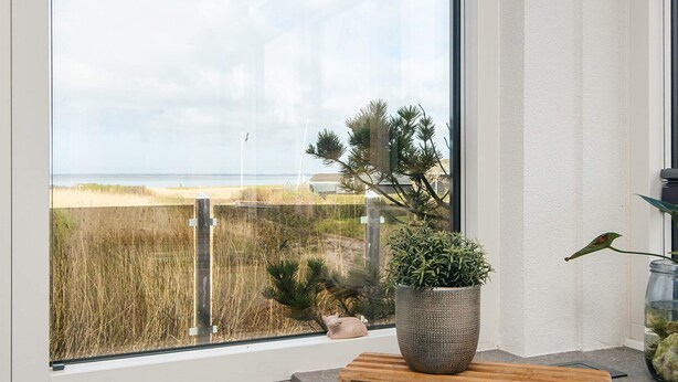 Eine Küche mit großem Fenster, das auf einen Strand und Dünen blickt, einem Holztisch zum Schneiden und einem Blumentopf.