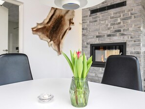 Ein Essbereich mit einem weißen Tisch, zwei schwarzen Stühlen, einer Glasvase mit grünen Blättern und einem Kamin vor einer Steinwand.