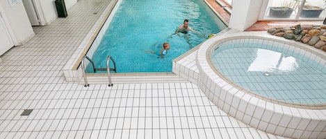 An indoor swimming pool with tiled flooring, a separate hot tub area, and a view of the outside.
