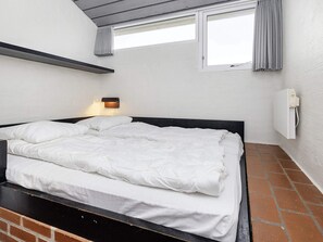 Una cama individual con ropa blanca en una habitación con paredes blancas, una ventana con cortinas y una lámpara fijada a la pared.