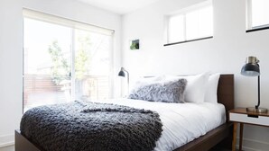 Ein modernes Schlafzimmer mit einem großen Bett, einem Nachttisch mit Lampe und einem Fenster mit Blick ins Freie.
