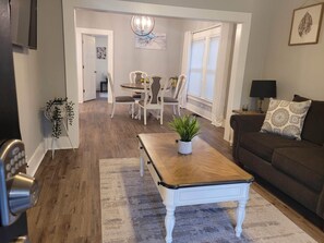A living room with a wooden coffee table, a brown sofa, and a white armchair. There is a dining area with a table and chairs in the background. - Charming 3b-1b Westside Indy (Indianapolis)