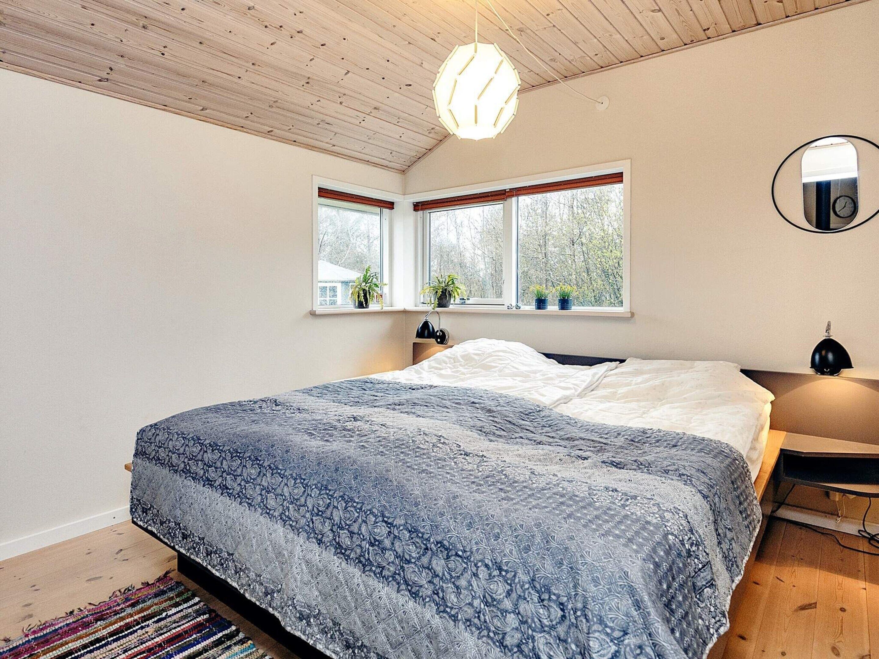 A bedroom with a large bed, two windows with blinds, a bedside table with a lamp, and a ceiling light fixture.