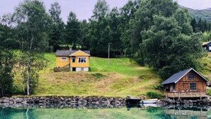 En hytte ved søen med et spejlbillede i vandet, omgivet af træer og en stenvæg.