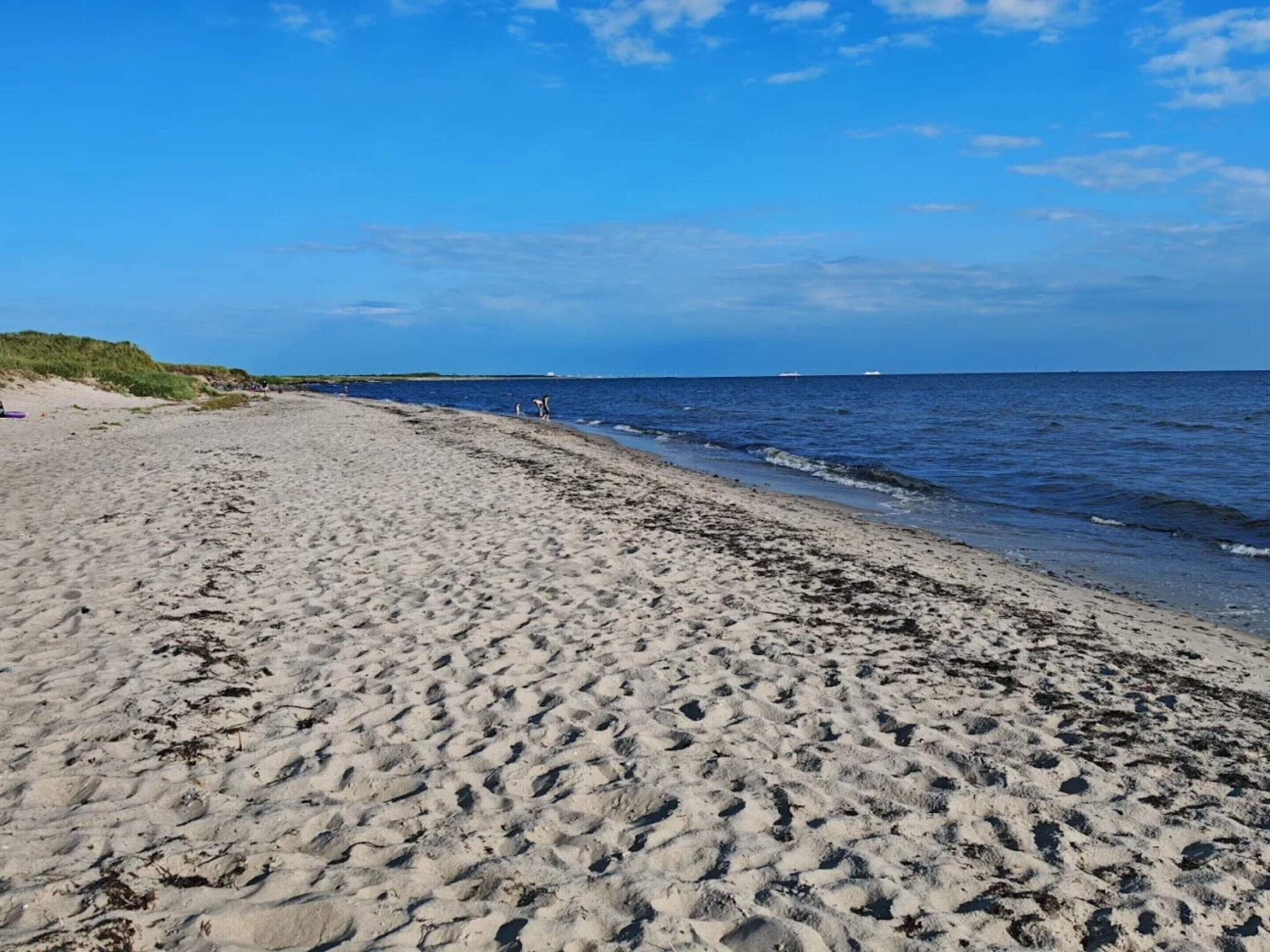 Une plage de sable fin aux vagues douces, un ciel dégagé et des bateaux à l’horizon.