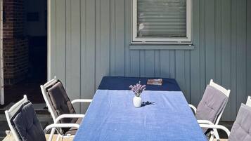 Ein überdachter Patio-Bereich mit Tisch und Stühlen, ein Fenster mit Jalousien und eine Ziegelwand.