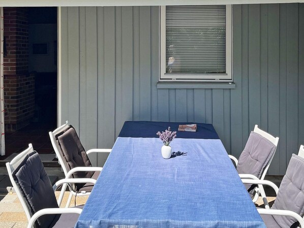A covered patio area with a table and chairs, a window with blinds, and a brick wall. - 10 Person Holiday Home in Dannemare (Dannemare)