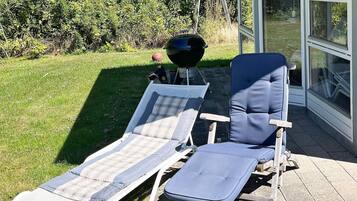 Zwei Liegestühle mit Kissen, ein Grill und eine Glastür, die in einen Garten führt.