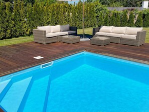 Una piscina con terraza de madera y dos sofás de mimbre con cojines.