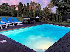 Una piscina rectangular con solárium de madera, rodeada de césped, árboles y una zona de barbacoa.