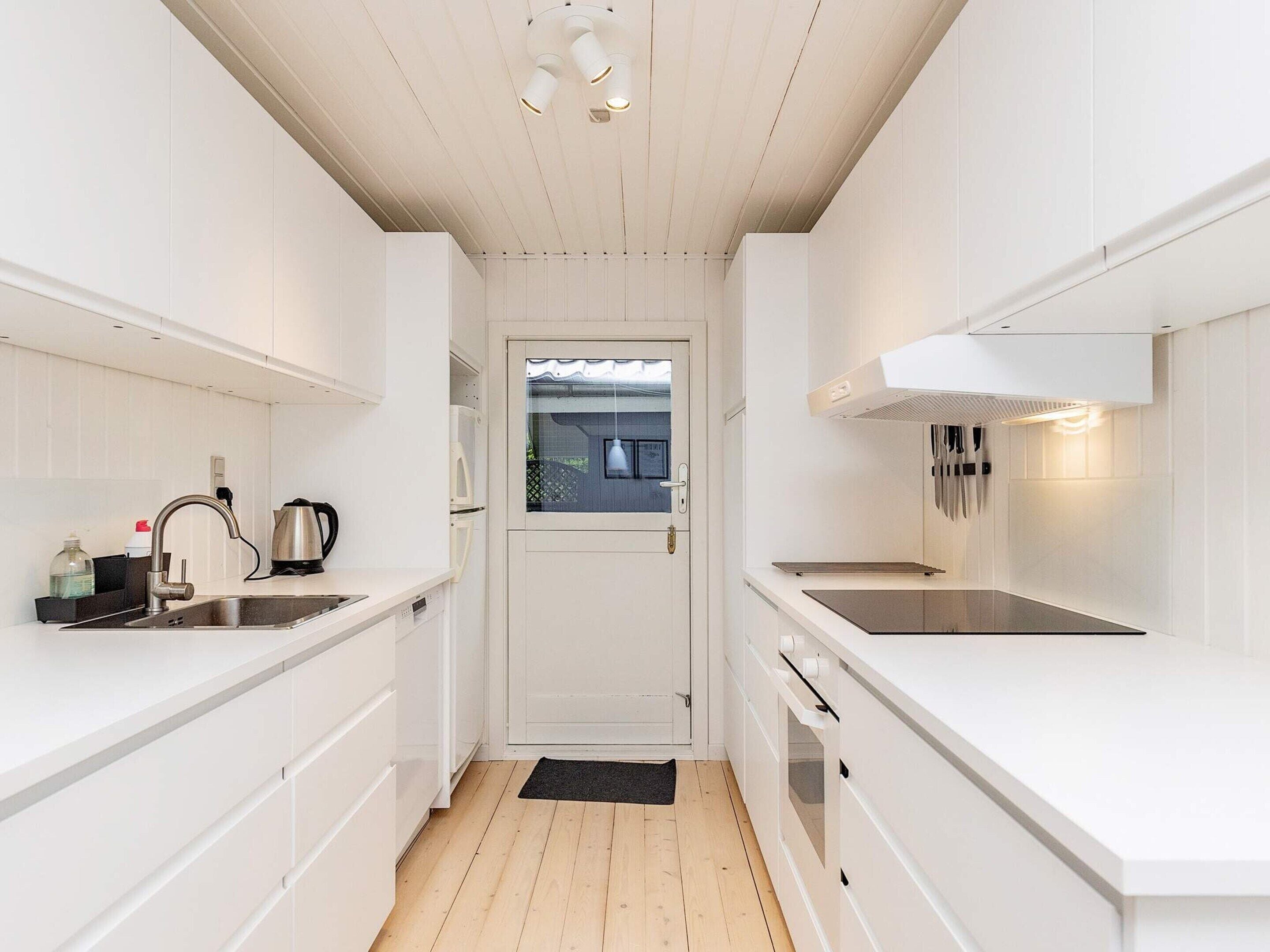 A narrow kitchen with white cabinetry, a stainless steel sink, a kettle, and a window with a view.