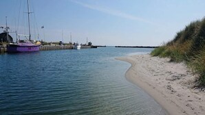 A sandy beach with a clear waterway, boats docked at a pier, and a grassy dune on the right.