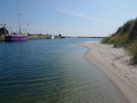 Una playa arenosa con una vía fluvial transparente, barcos amarrados en un muelle y una duna cubierta de hierba a la derecha.