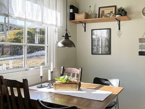 Un coin repas comprenant une table, des chaises, une fenêtre avec des rideaux, un luminaire suspendu et une étagère murale agrémentée d’objets décoratifs.