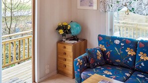 Quarto com piso de madeira, sofá floral azul, mesa lateral de madeira, cômoda de madeira com um globo e flores, e varanda com guarda-corrimão.