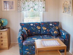A room with a floral-patterned sofa, a wooden side table with a globe, and a window with a lace curtain. - 4 Person Holiday Home in Hallevadsholm (Hällevadsholm)