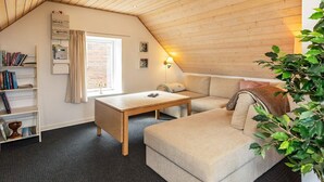 A cozy attic room with a wooden ceiling, a window with curtains, a bookshelf, a sofa, and a small table.