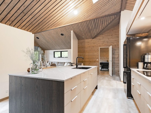 A modern kitchen with a large island, wooden ceiling, and a view to the living area.