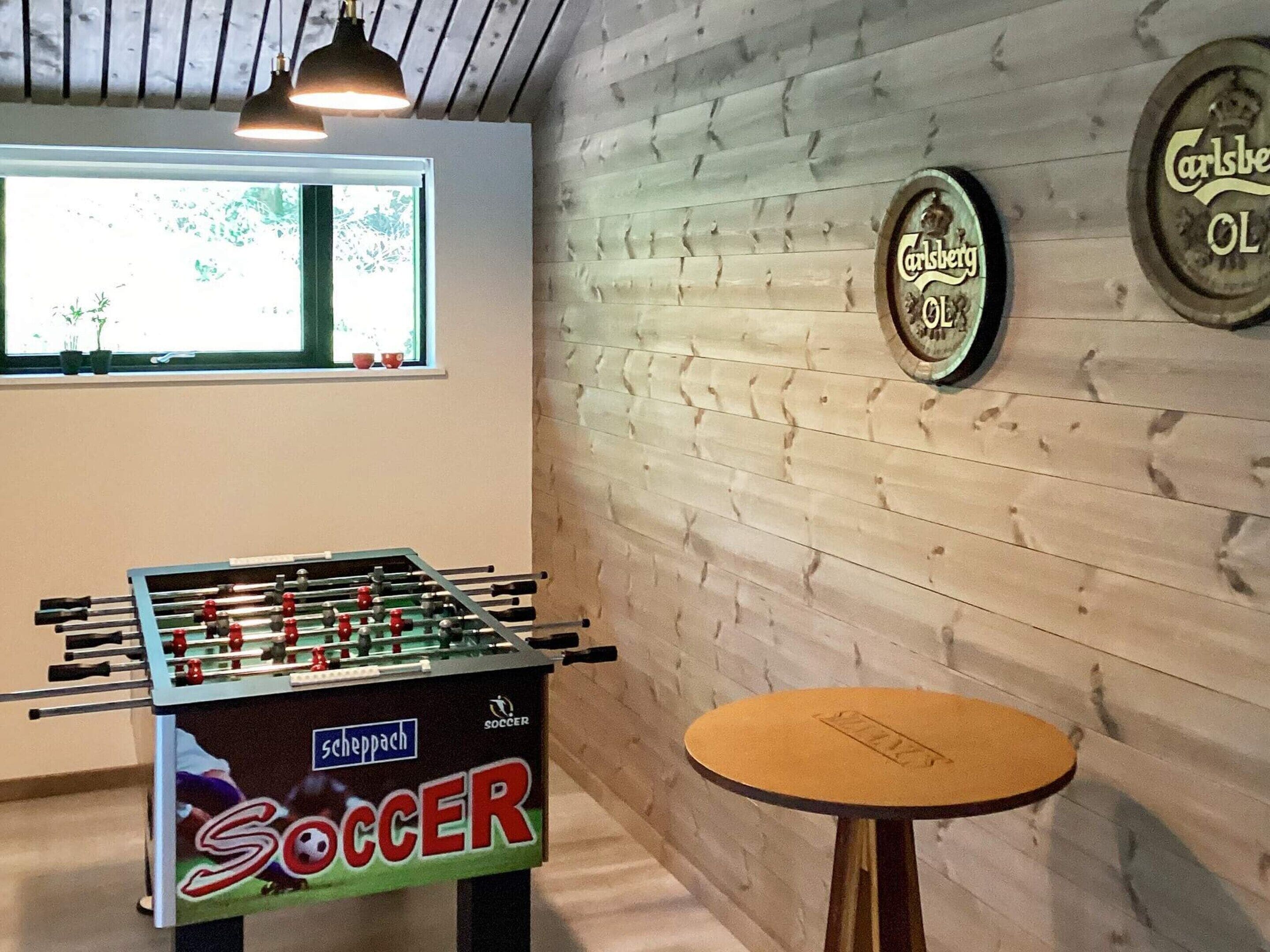 Et bordtennisbord med teksten 'fodbold' og et rundt bord med et logo, i et rum med trævægge og et vindue.