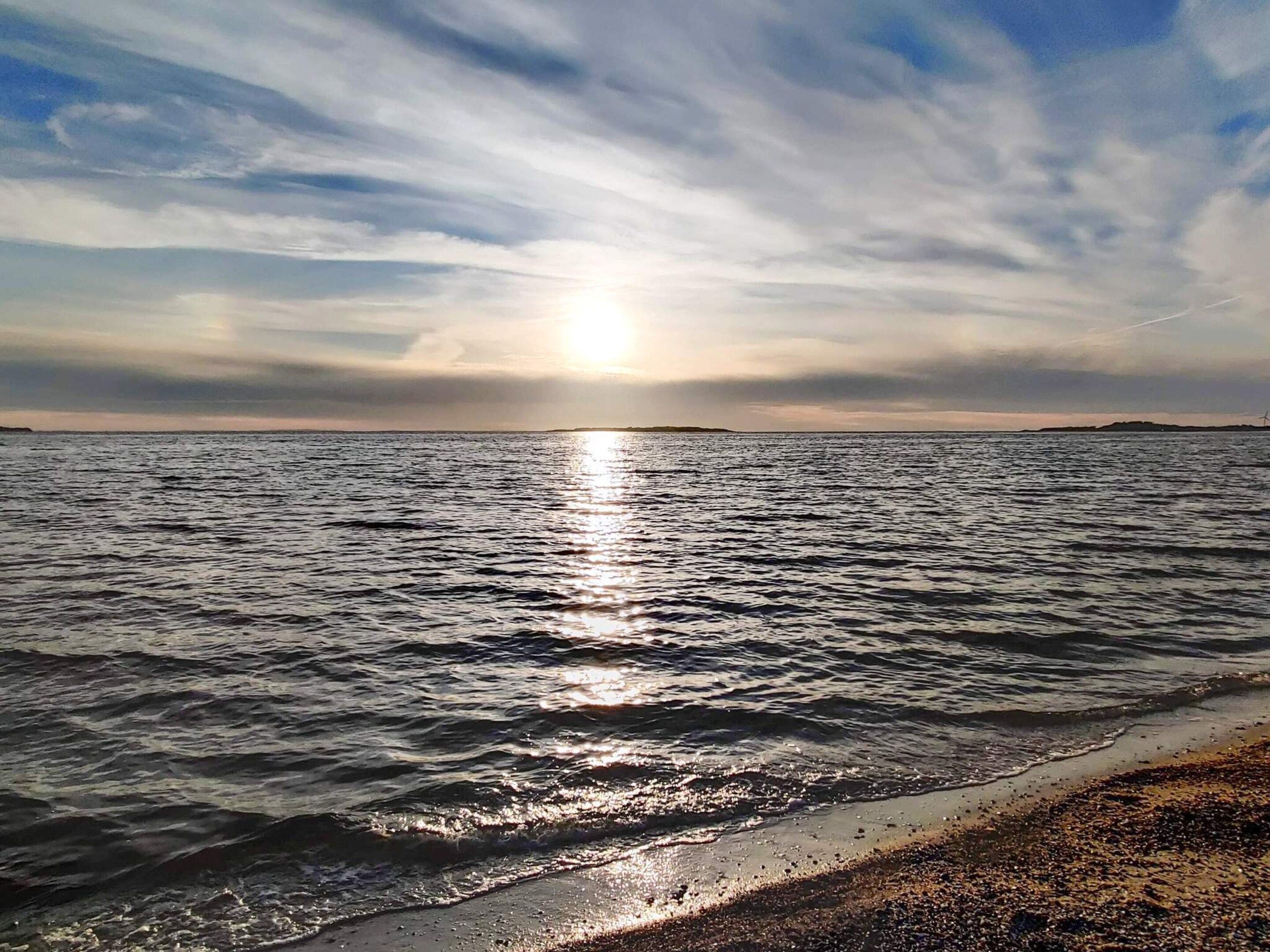 Una playa con agua que refleja la luz del sol, una masa de tierra distante y un cielo nublado.
