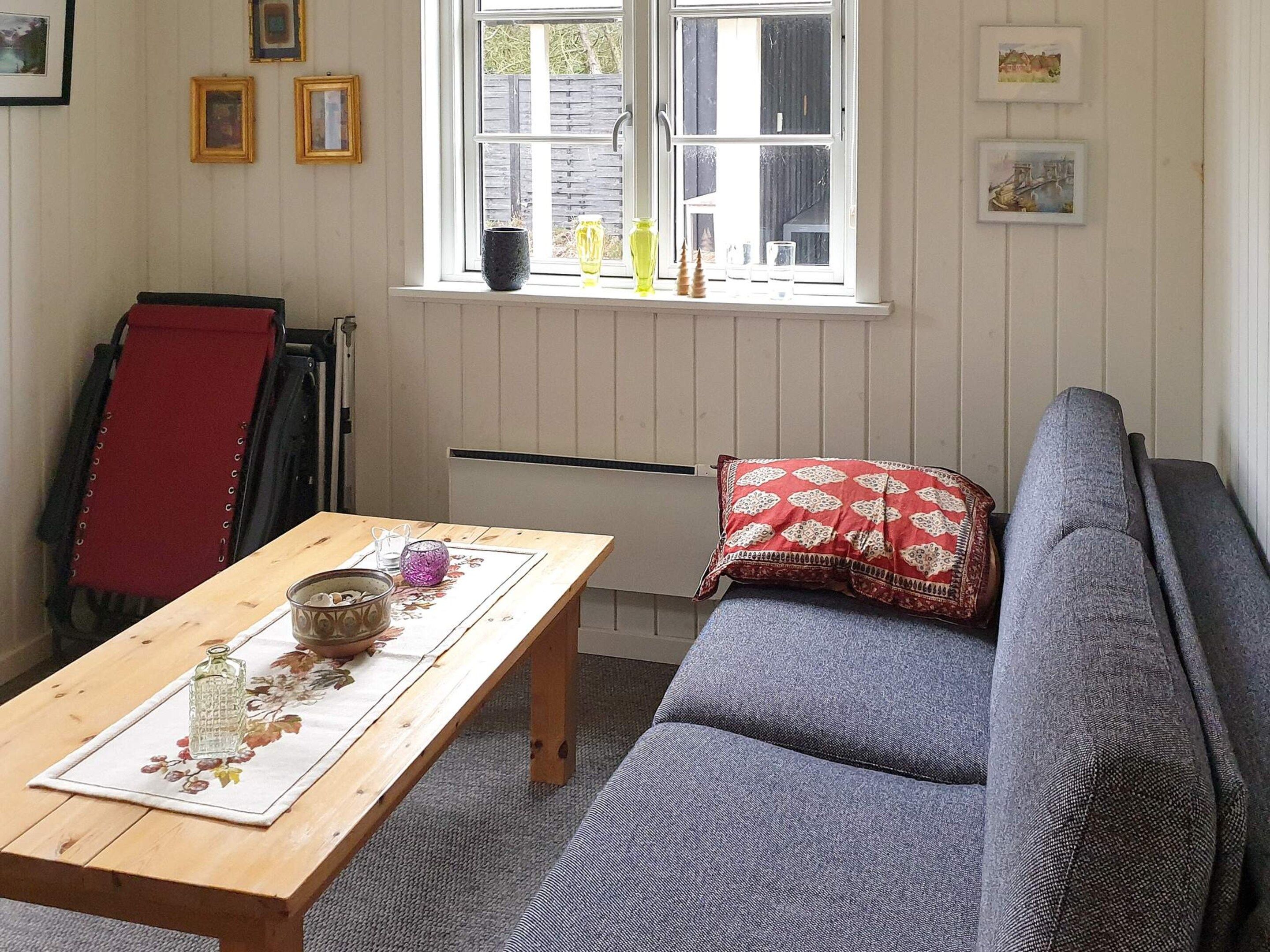 Una habitación con una mesa de madera, un sofá gris, un cojín rojo y una ventana con cuadros en la pared.