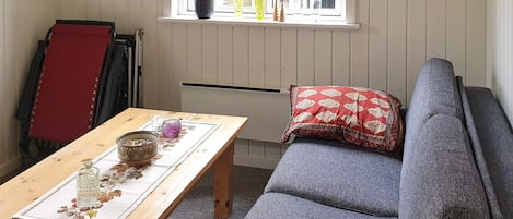 Una habitación con una mesa de madera, un sofá gris, un cojín rojo y una ventana con cuadros en la pared.