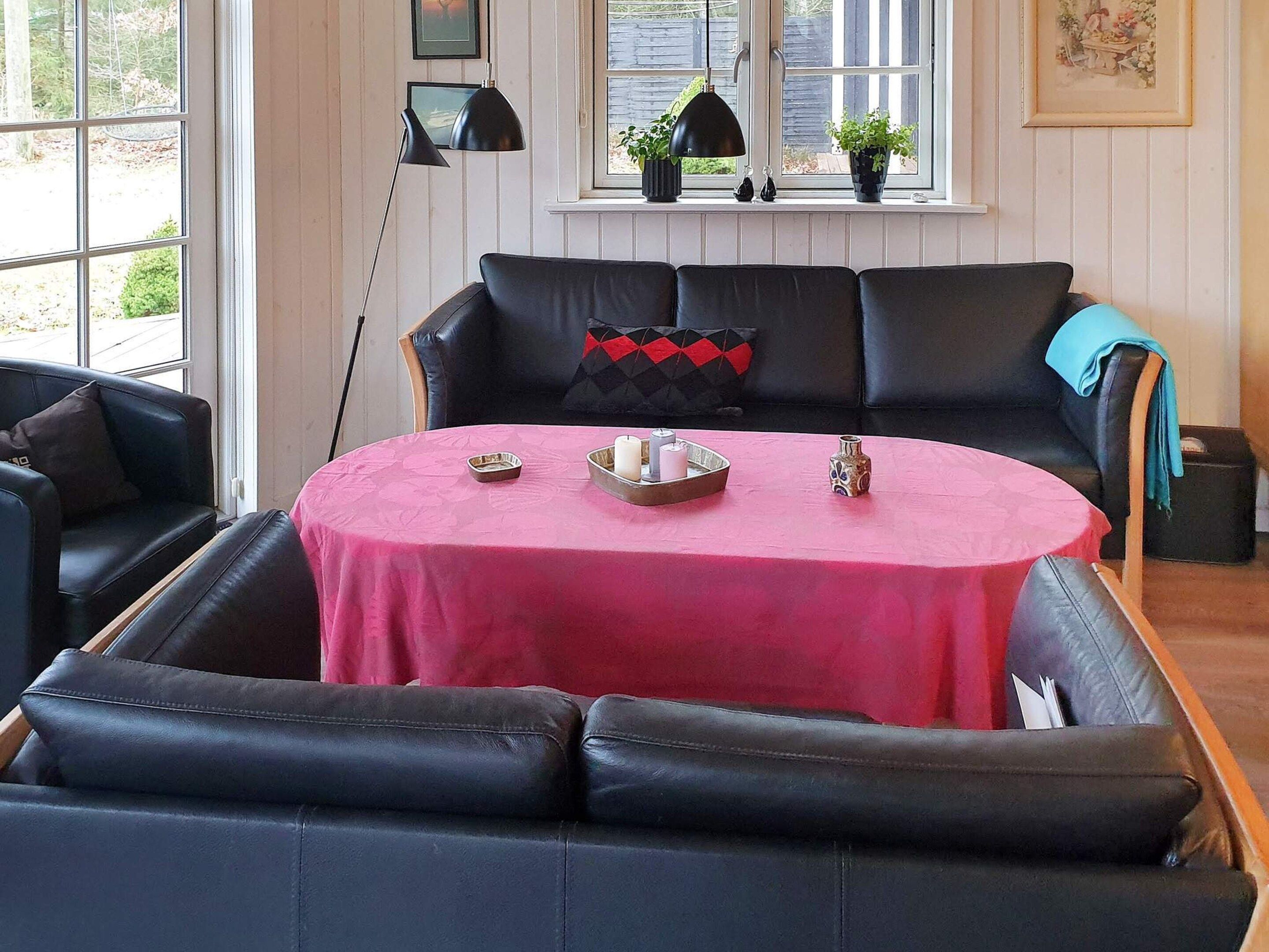 Una habitación con un sofá de cuero negro, una mesa redonda con mantel rosa y una ventana con plantas en macetas.