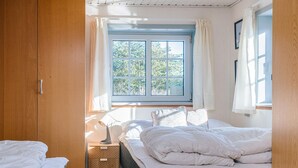 Une chambre avec deux lits, une fenêtre avec des rideaux, une table de chevet avec une lampe, et une vue sur la verdure à l’extérieur.