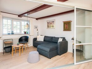Un salon moderne avec un canapé d’angle gris, une table ronde en bois, une chaise noire et un pouf gris. On y trouve également un radiateur, une fenêtre avec des rideaux et un tableau encadré accroché au mur.