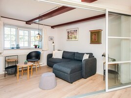 A modern living room with a grey sectional sofa, a round wooden table, a black chair, and a grey ottoman. There is a radiator, a window with curtains, and a framed picture on the wall.