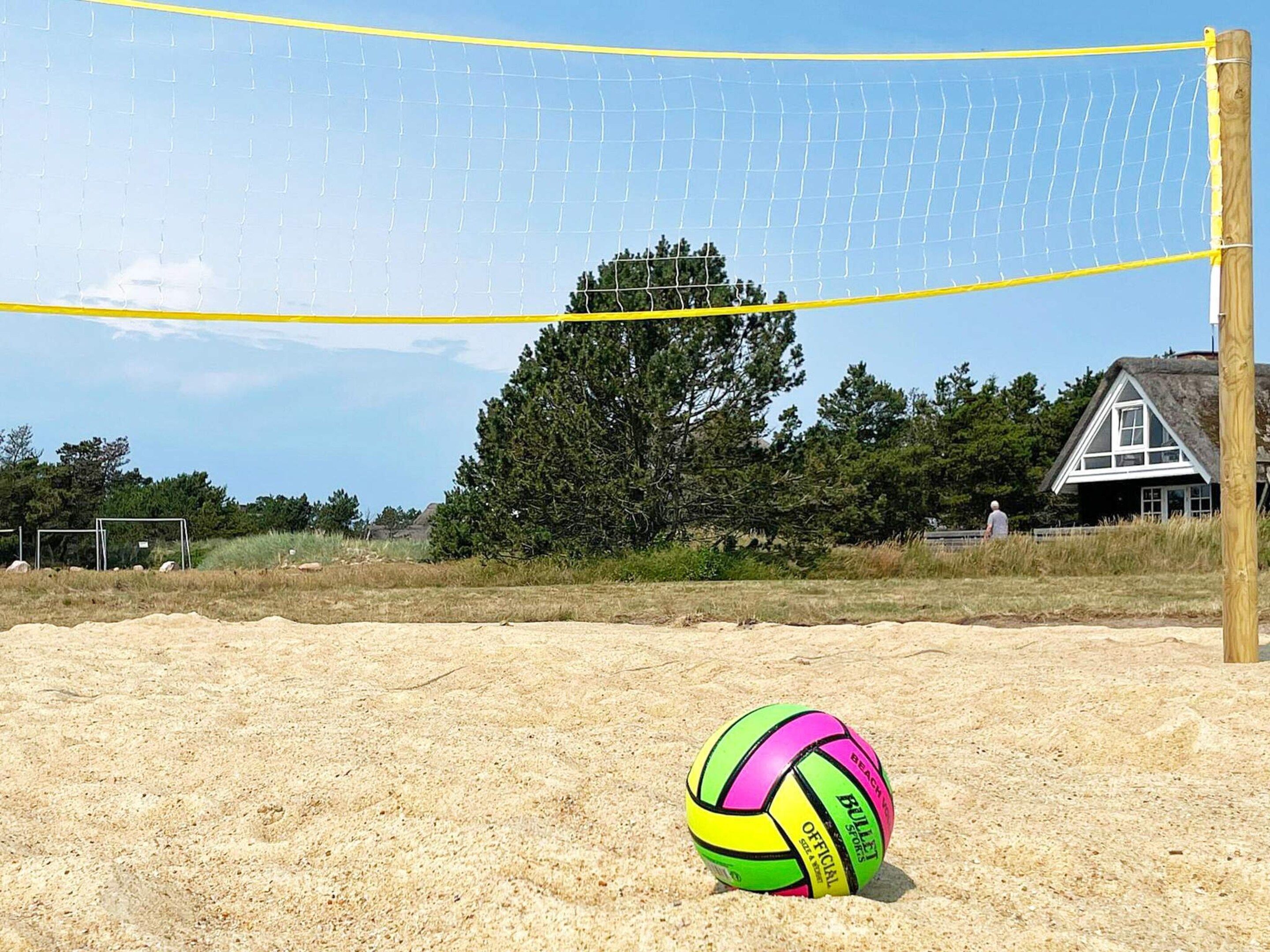 A beach volleyball net with a ball on the sand, a wooden post, and a house in the background.