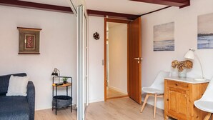 Un salon moderne avec un canapé gris, une petite table d’appoint avec une lampe, un meuble en bois et une porte donnant sur une autre pièce.