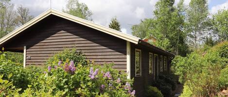 A wooden cabin with a sloped roof, surrounded by lush greenery and a clear sky.