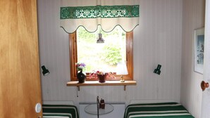 A room with two single beds, a window with a green lace curtain, a small table with a lamp, and a door.