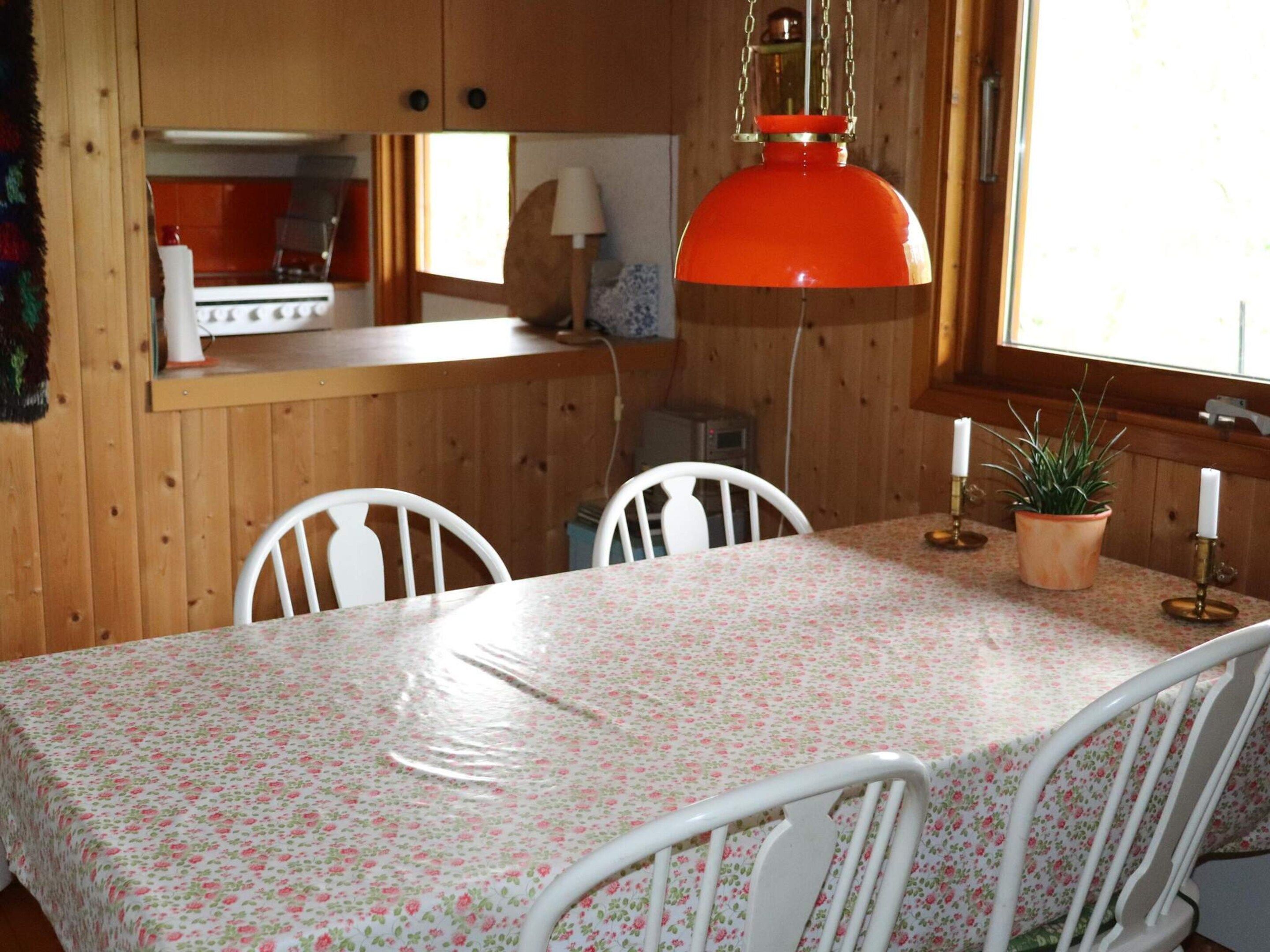 A dining area with a table covered in a floral tablecloth, white chairs, a hanging orange pendant light, and a window with a view.
