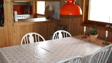 A dining area with a table covered in a floral tablecloth, white chairs, a hanging orange pendant light, and a window with a view.