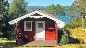 A small red cabin with a white door and windows, surrounded by trees and a lake. - 7 Person Holiday Home in Tyreso-by Traum (Tyreso)