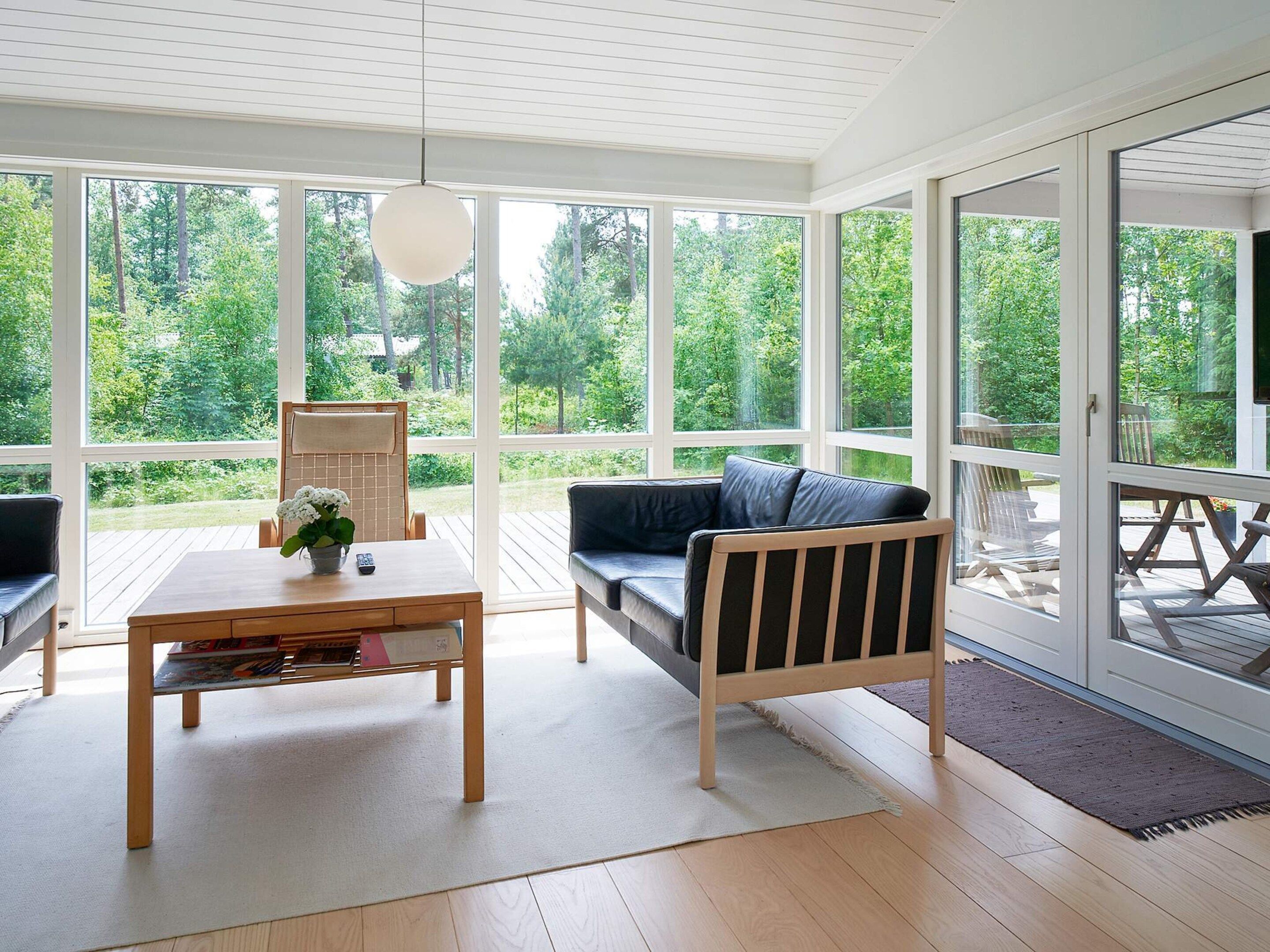 Ein sonnendurchflutetes Zimmer mit einem Holztisch, einer Couch und Blick auf eine Terrasse und grüne Bepflanzung.