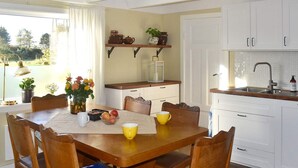 Ein Essbereich mit einem Holztisch, sechs Stühlen und einem Blumenstrauß. Es gibt eine Küchenzeile mit weißen Schränken und einem Spülbecken.