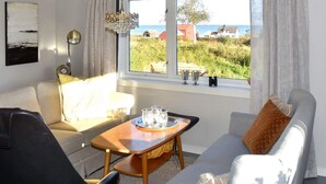Ein Wohnzimmer mit einer Couch, einem Sessel, einem kleinen Tisch mit Gläsern und Meerblick durch ein Fenster.